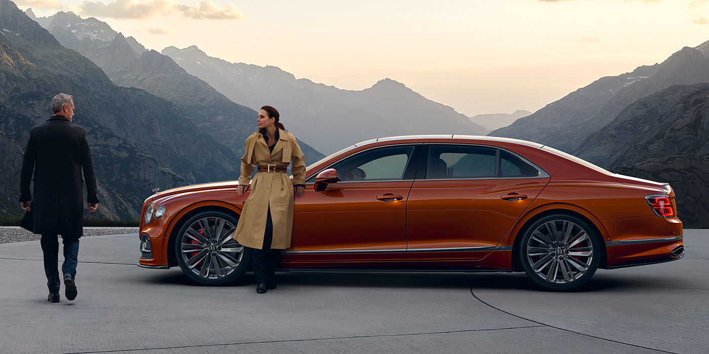 Bentley Milano Bentley Flying Spur Speed parked in Orange Flame coloured exterior parked, with mountainous background and two people in view.