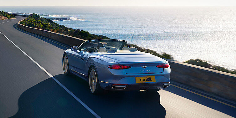 Bentley Milano New Bentley Continental GTC Azure convertible rear view in Light Windsor blue paint with 22 inch ten spoke wheels driving dynamically on an ocean road in the sunshine